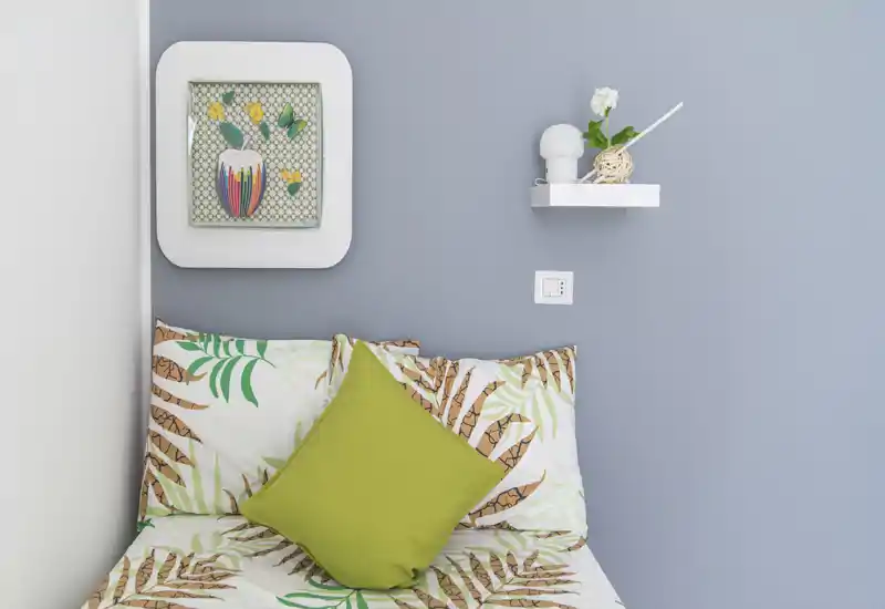 Bright, neatly styled bedroom corner with patterned bedding and a green accent cushion; a floating shelf and wall art add tasteful decor.
