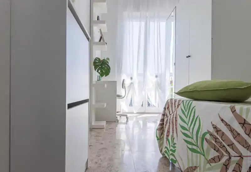 Bright modern bedroom with a leafy-patterned bedspread, green cushion and sheer curtains that let in abundant natural light. Clean white furnishings and a small desk area create a fresh, airy atmosphere ideal for rentals.