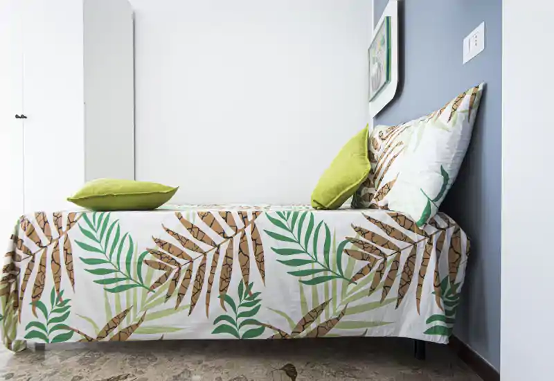 Bright, modern single bedroom with patterned tropical bedding and green accent pillows; clean white walls and a simple wardrobe create a fresh, welcoming space.
