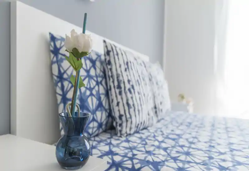 Camera da letto luminosa e curata con biancheria blu a motivi e cuscini coordinati; un piccolo vaso sul comodino aggiunge un tocco decorativo e luce naturale soffusa.