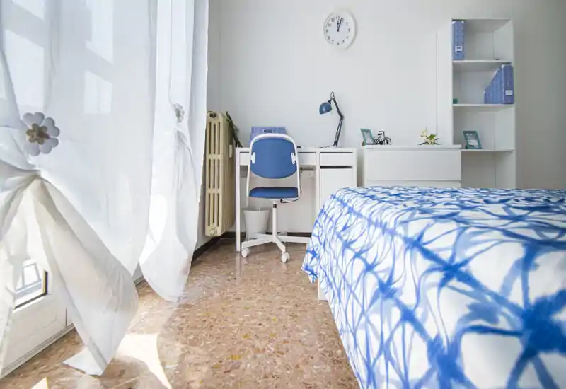 Dormitorio luminoso y ordenado con colcha azul y blanca, escritorio y silla junto a una ventana soleada — ideal para una persona o estudiante.