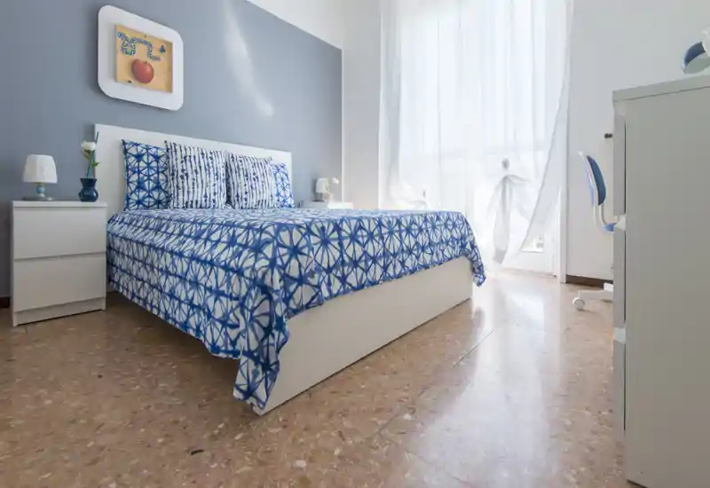 Dormitorio luminoso y bien presentado con cama blanca, ropa de cama azul estampada, mesitas de noche y luz natural de una gran ventana.