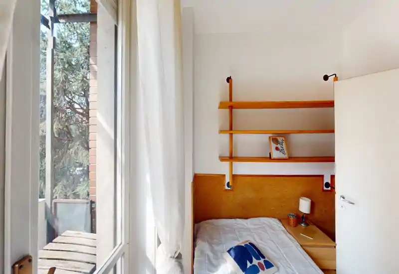 Dormitorio compacto y luminoso con cama individual, estanterías de madera y mesita; la luz del balcón crea un ambiente cálido y acogedor.