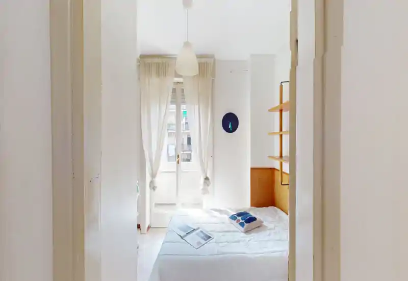 Dormitorio luminoso y minimalista con una cama doble bien hecha, ropa de cama blanca, un cojín decorativo y estanterías a la derecha; cortinas semitransparentes dejan entrar abundante luz natural desde la puerta al balcón.