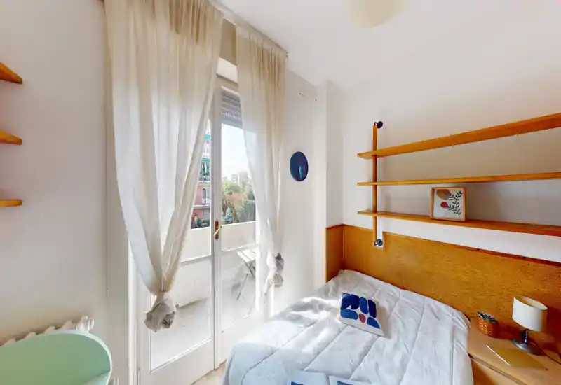 Dormitorio pequeño y luminoso con cama doble junto a una cabecera de madera, puertas al balcón con cortinas ligeras y estanterías empotradas — acogedor y perfecto para estancias en la ciudad.