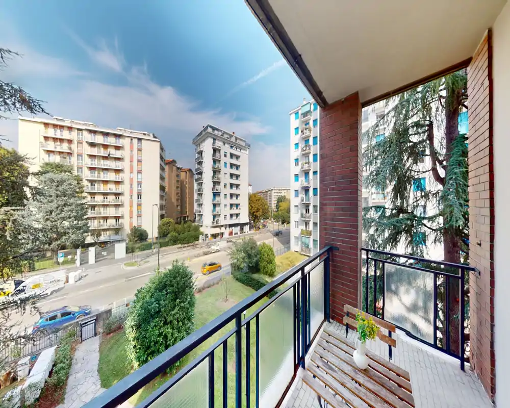 Balcone soleggiato con una panca in legno e una pianta in vaso, con vista su una strada alberata e palazzi residenziali.