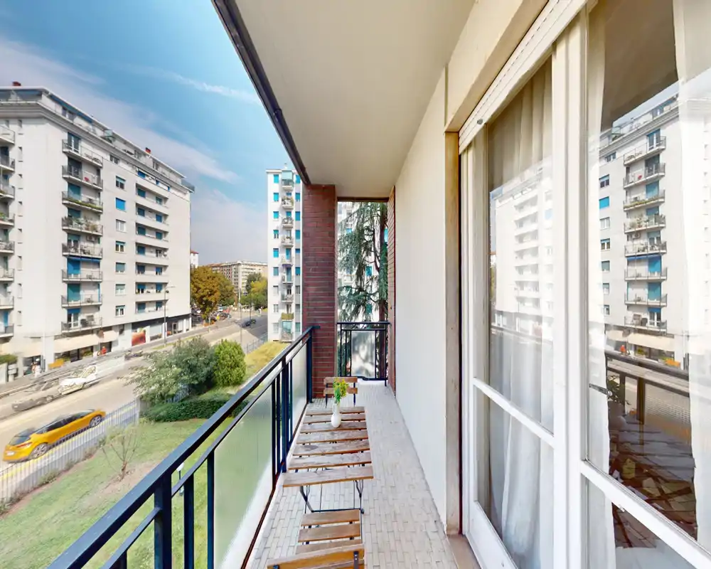 Balcone soleggiato e stretto con un piccolo tavolo e sedie in legno, affaccia su una strada alberata e palazzi vicini — perfetto per il caffè mattutino.