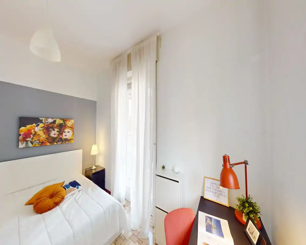 Camera da letto luminosa e ben arredata con letto bianco, cuscini decorativi, lampada da comodino e una piccola scrivania vicino alla finestra — tende leggere che lasciano passare la luce naturale.
