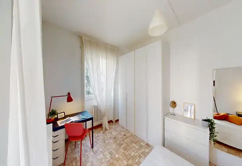 Camera da letto luminosa e ben allestita con armadio bianco, comò e una piccola scrivania vicino alla finestra. Luce naturale e dettagli caldi rendono l'ambiente accogliente e adatto agli affitti brevi.