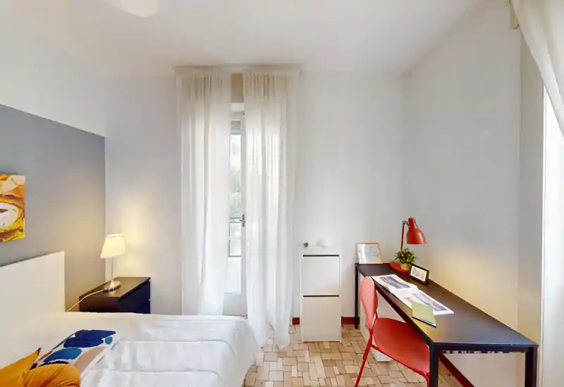Camera da letto luminosa e ordinata con letto matrimoniale comodo, comodino con lampada e una zona studio con scrivania e sedia vicino alla finestra con tenda leggera che lascia entrare luce naturale.