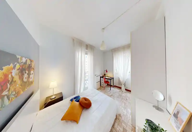 Camera da letto luminosa e ben composta con letto matrimoniale, cuscini decorativi e una lampada da comodino. La luce naturale filtra attraverso le tende leggere e una piccola scrivania vicino alla finestra crea un angolo lavoro accogliente.