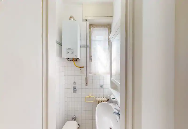 Small, bright bathroom with wall-mounted boiler, tiled walls, sink and toilet visible; compact and well-lit space suitable for rental listing photos.