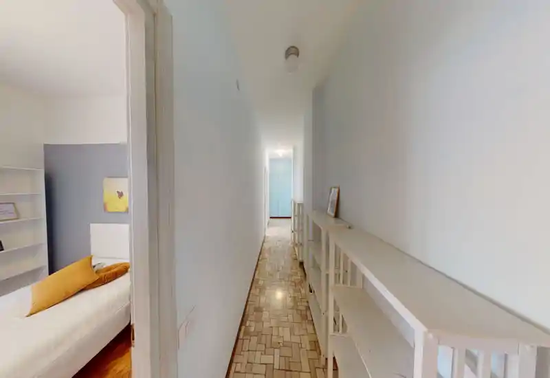 Dormitorio compacto visto desde un pasillo estrecho: cama individual con cojines y decoración sutil, estanterías blancas y cojines cálidos. Espacio luminoso y funcional, ideal para alquileres temporales o estudiantes.