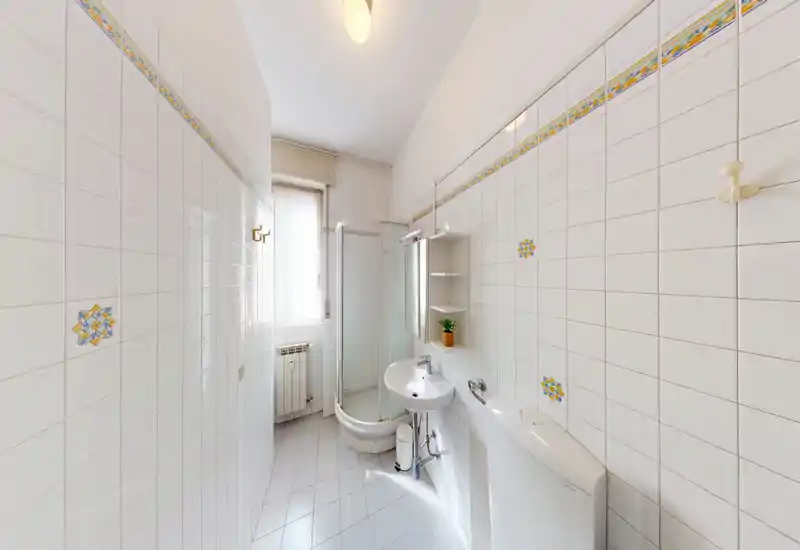 Bright, tidy bathroom with corner shower, wall-mounted sink and toilet; white tiles with decorative accents and natural light from a window.