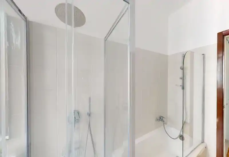Bright, modern bathroom with a combined bathtub and glass shower enclosure, wall-mounted showerhead and handheld fixture, and neutral tile walls.