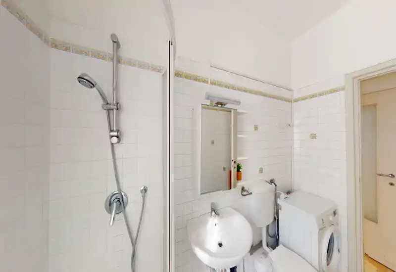 Compact tiled bathroom with a shower cabin, wall-mounted sink, mirror cabinet and a washing machine; bright, clean and well-lit space suitable for apartment listings.