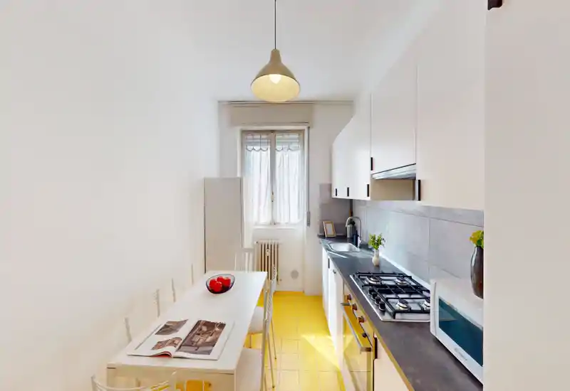 Bright, modern galley kitchen with white cabinets, dark countertop, gas hob and a small dining table; yellow tiled floor and natural light from the window create a cheerful atmosphere.