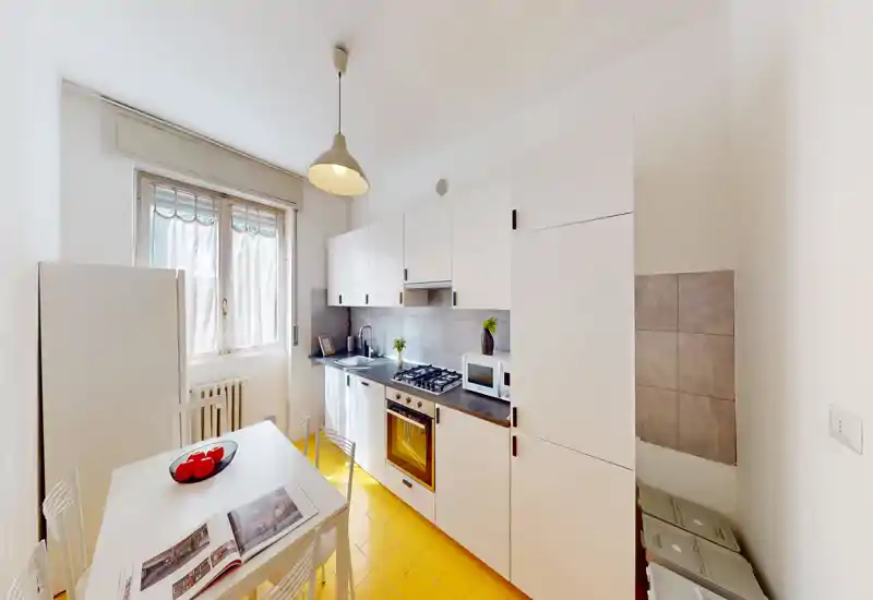Bright, compact kitchen with white cabinets, built-in oven and gas hob, a small dining table and large window bringing natural light.