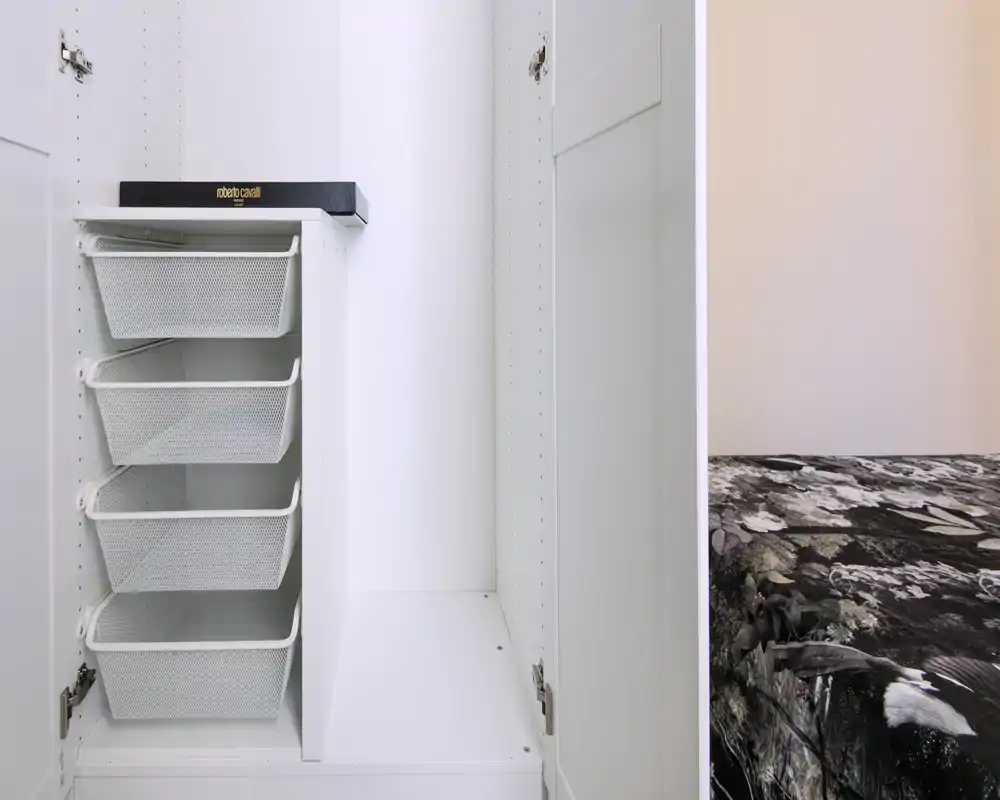 A bright bedroom view showing an open white wardrobe with mesh storage drawers and a partial view of the bed with patterned bedding at the right edge.