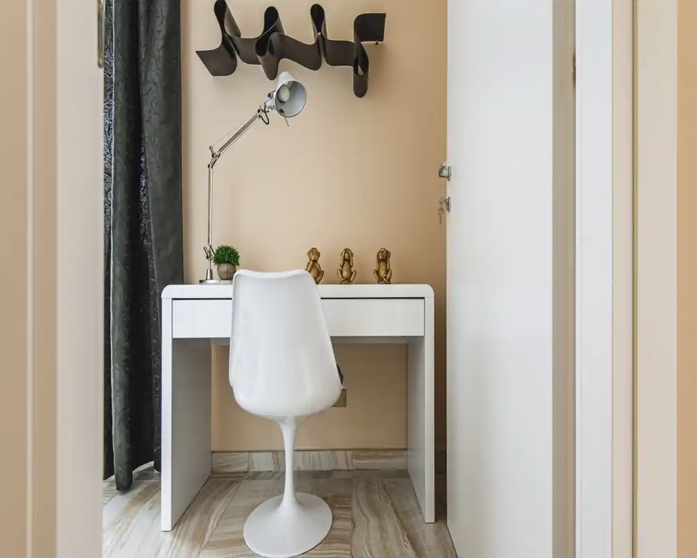 A compact home office nook within a living space: white desk with a modern swivel chair, adjustable desk lamp, decorative figurines and a small plant against a warm beige wall and heavy curtains.