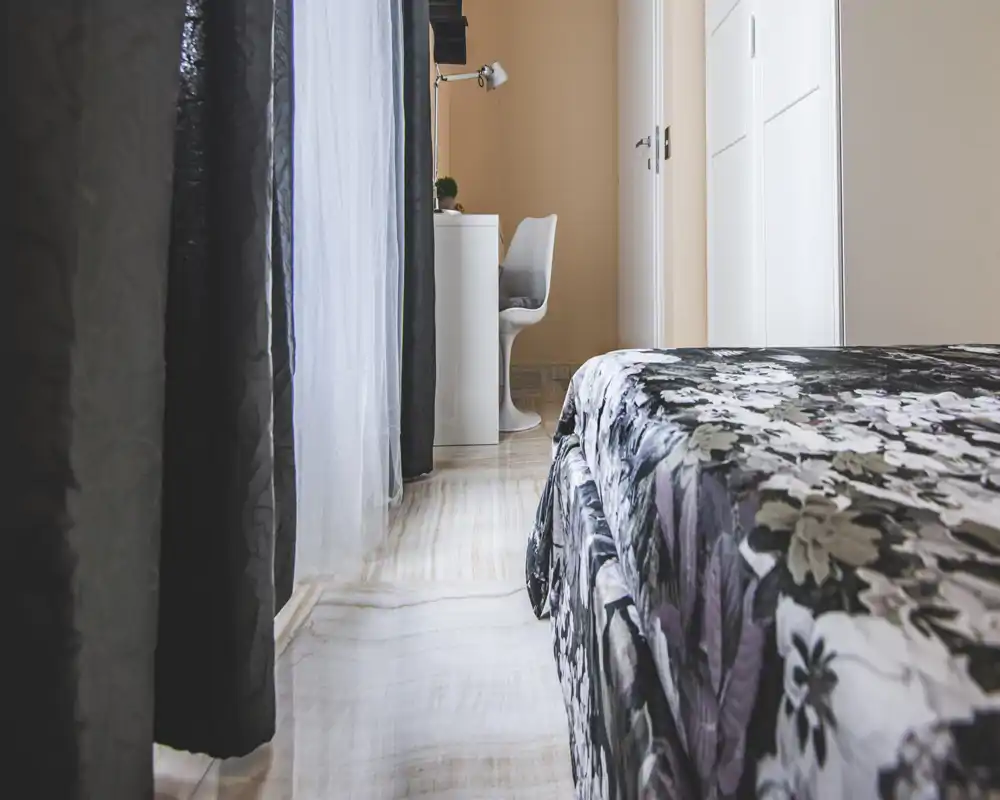 Bright, modern bedroom view with a patterned bedspread, floor-to-ceiling curtains and a small desk area by the window — a cozy, well-lit sleeping corner.