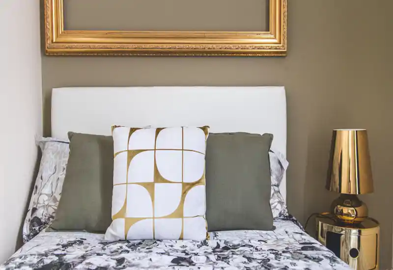 Stylish bedroom corner with a white upholstered headboard, decorative pillows and patterned bedding; warm gold accents and a bedside lamp add a cozy, modern touch.
