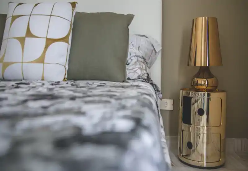 Close-up of a stylish bedroom corner featuring pillows, patterned bedding and a reflective gold bedside table with lamp. The image highlights texture and warm tones for a cozy atmosphere.