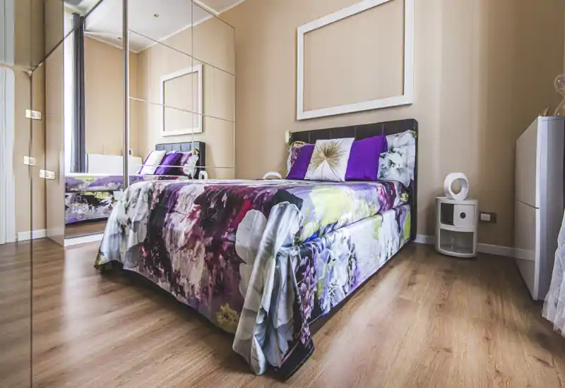Bright, stylish bedroom with a large bed dressed in a colorful floral duvet, decorative pillows and mirrored wardrobe reflecting depth. Warm wooden floors and neutral walls create a cozy, contemporary atmosphere ideal for rental photos.