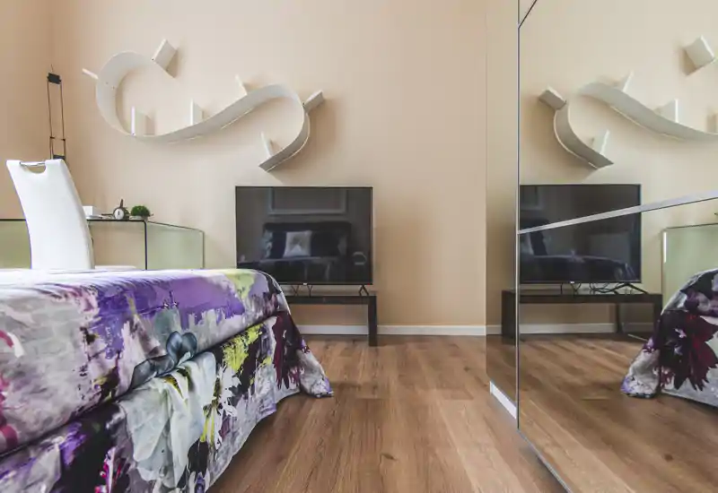 Camera da letto moderna e luminosa con copriletto fantasia, mobile TV basso e armadio con specchio che amplia lo spazio. Toni caldi e mensole decorative creano un'atmosfera accogliente, perfetta per annunci.