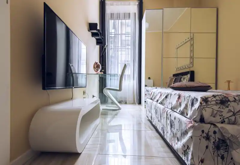 Bright, stylish bedroom with patterned bedding, a mirrored wardrobe and a sleek glass desk by the window. Polished floors and tasteful lighting create a modern, welcoming atmosphere ideal for rental photos.