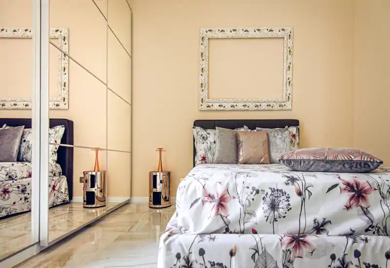 Bright, stylish bedroom with floral bedding, decorative cushions and a mirrored wardrobe that enlarges the space. Warm tones and reflective surfaces create an elegant, cozy atmosphere.