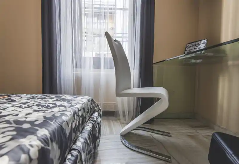 Angolo di camera moderna con copriletto a fantasia, tende leggere e una sedia bianca dal design elegante vicino a una scrivania in vetro. La luce naturale dalla finestra illumina lo spazio pulito e minimal.