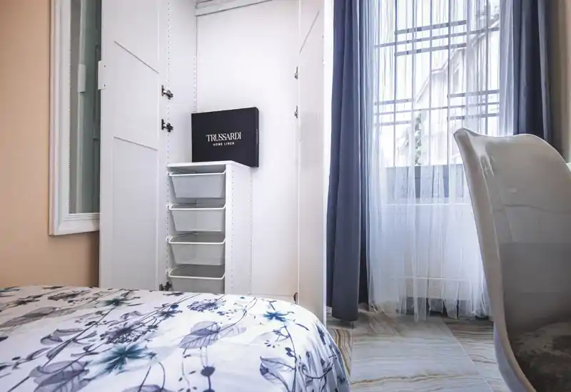 Bright, neatly styled bedroom corner featuring a floral-patterned bedspread, built-in wardrobe with storage baskets and a window with sheer curtains that fills the space with natural light.