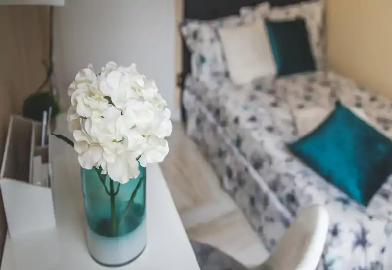 Bright, stylish single bedroom corner with patterned bedding and teal accent cushions; a vase with white flowers on a desk adds a fresh, decorative touch.
