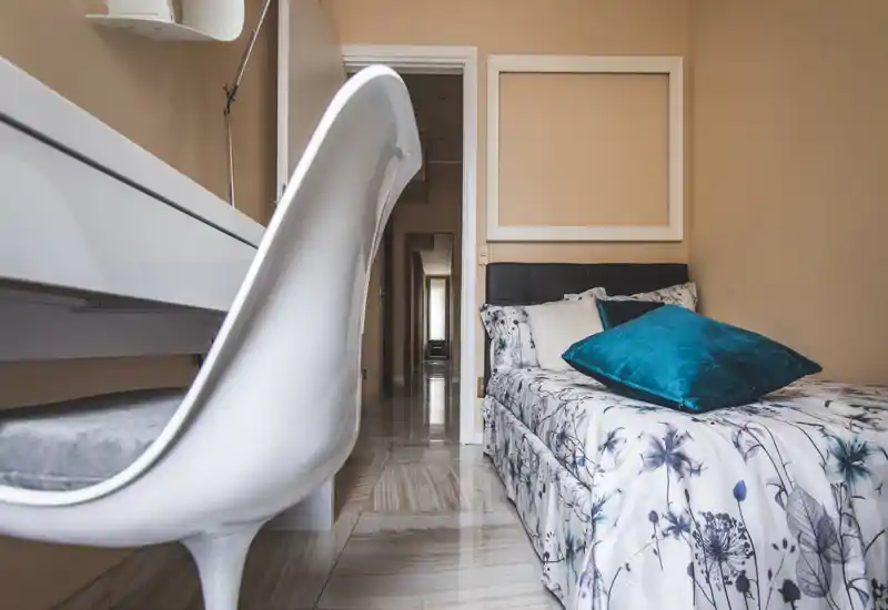 Neat single bedroom with patterned bedding and a plush teal cushion; a modern white desk chair in the foreground adds depth and a workspace feel.