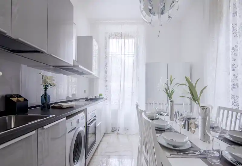 Bright, modern kitchen with integrated appliances and a set dining table; airy white finishes and decorative flowers create a stylish, inviting space.