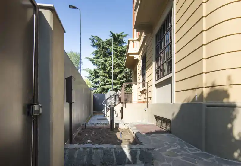 Sunny side yard / narrow outdoor passage beside the building, with paved walkway, small planting bed and metal gate — suitable to show outdoor access and building exterior.