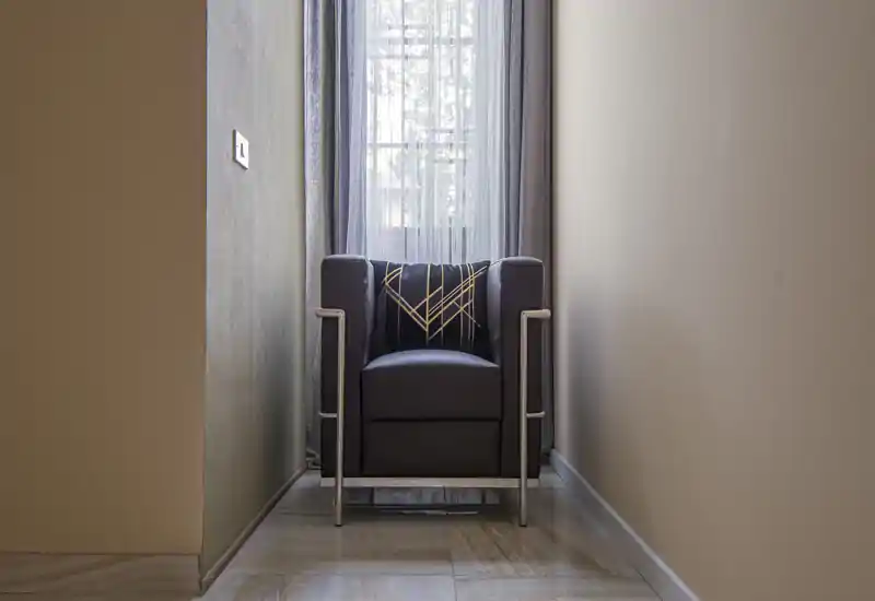 A modern accent armchair is placed at the end of a narrow corridor in front of a sheer window curtain, creating a quiet reading nook with a decorative cushion.