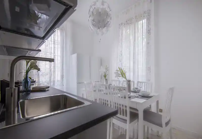 Bright, all-white kitchen with a black countertop, stainless sink and a dining table set for guests; tall windows with sheer curtains fill the room with natural light.