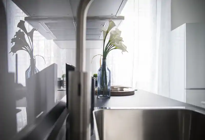 Bright modern kitchen countertop with a stainless sink in the foreground and a decorative vase with calla lilies on the work surface; soft natural light and minimal, stylish styling.