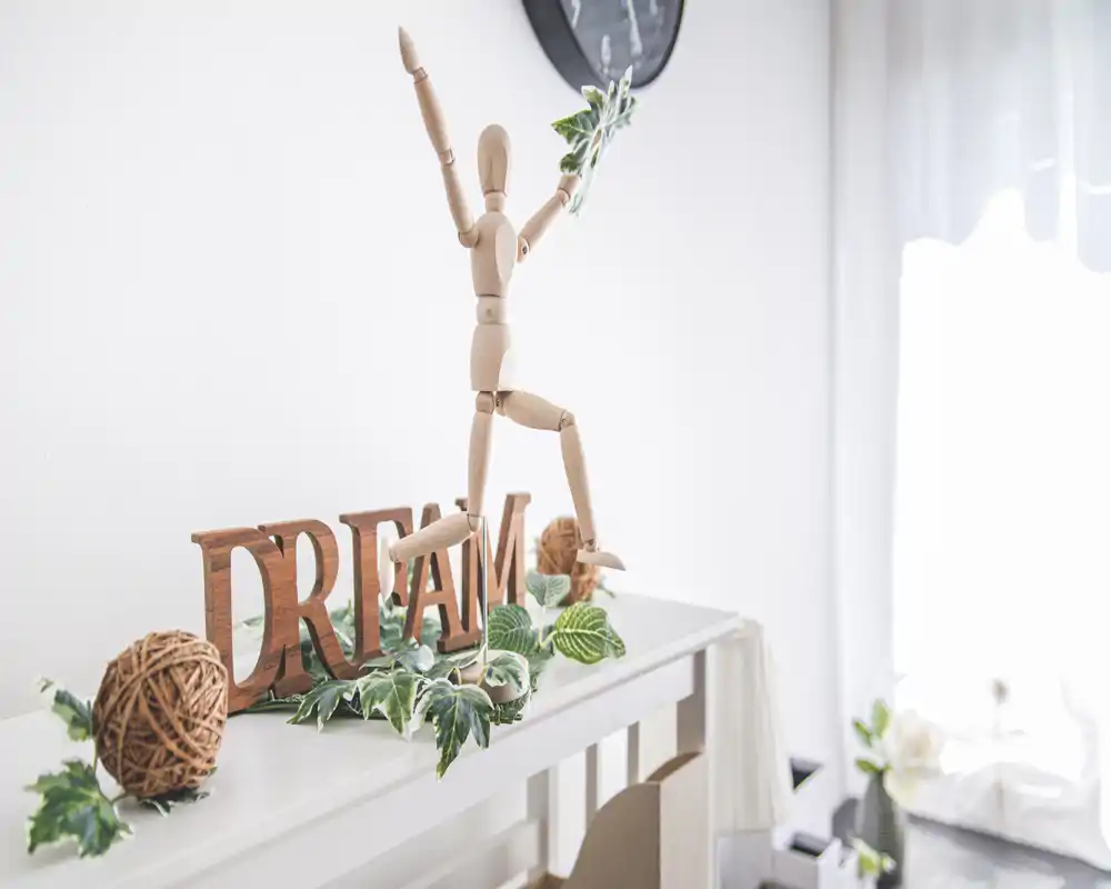 Angolo d'ingresso luminoso e minimale con fogliame decorativo, insegna in legno 'DREAM' e un manichino in legno su una consolle bianca vicino a una finestra luminosa.