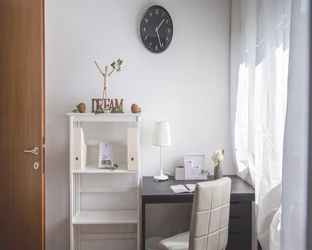 Angolo studio luminoso e minimal con scrivania, sedia, lampada e mensole decorative accanto a una finestra soleggiata.