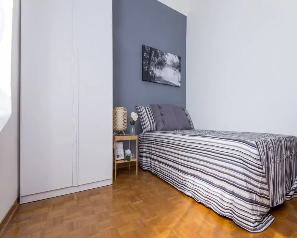 Camera singola luminosa e ben curata con biancheria a righe, comodino e armadio. Toni neutri e un quadro paesaggistico creano un'atmosfera rilassante, perfetta per un annuncio.