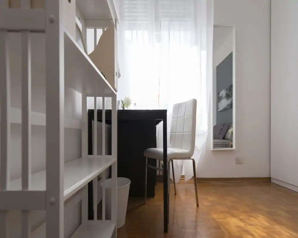 Angolo camera compatta con una piccola scrivania e sedia vicino a una finestra luminosa; il letto è visibile solo come riflesso nello specchio a parete. Lo spazio è ordinato e minimal, adatto per una persona.