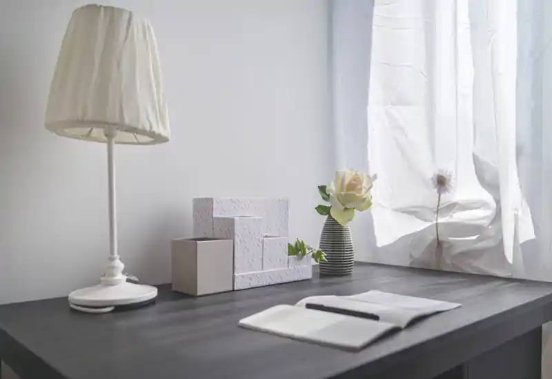 Coin bureau lumineux et minimaliste avec une lampe blanche, des boîtes décoratives, un petit vase contenant une rose et un carnet ouvert près d'une fenêtre baignée de lumière avec des rideaux légers.