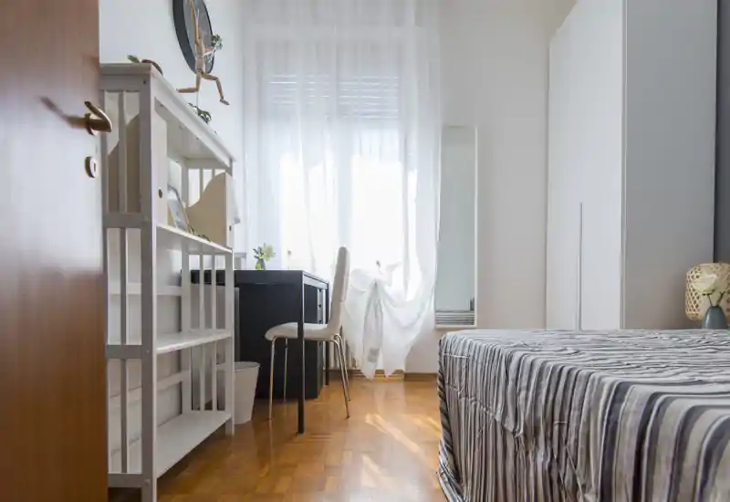 Camera da letto luminosa e ordinata con copriletto a righe, scrivania davanti a una finestra con tenda leggera, armadio e scaffalatura — perfetta per un soggiorno singolo o doppio confortevole.