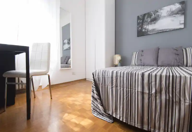 Camera da letto luminosa e dallo stile minimale con copriletto a righe, cuscini decorativi e luce naturale da una grande finestra; è presente una scrivania con sedia accanto al letto.