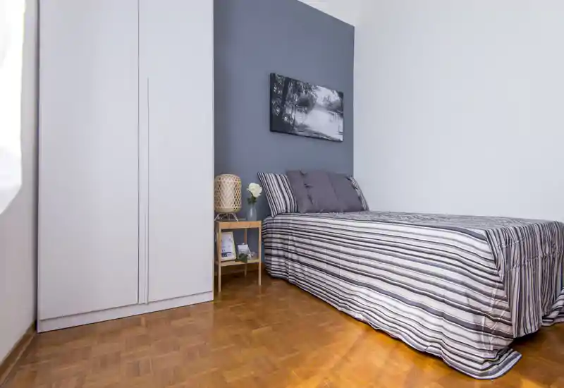 Camera singola luminosa e ben curata con biancheria a righe, comodino e armadio. Toni neutri e un quadro paesaggistico creano un'atmosfera rilassante, perfetta per un annuncio.