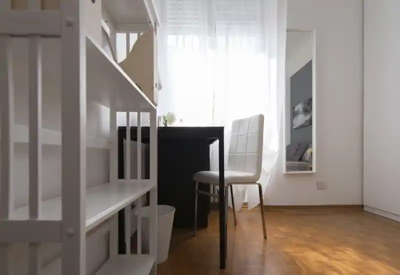Coin de chambre minimaliste et lumineux avec un bureau et une chaise ; le lit n’est visible que dans le reflet du miroir pleine longueur. La lumière naturelle traverse les voilages, offrant une atmosphère propre et aérée.