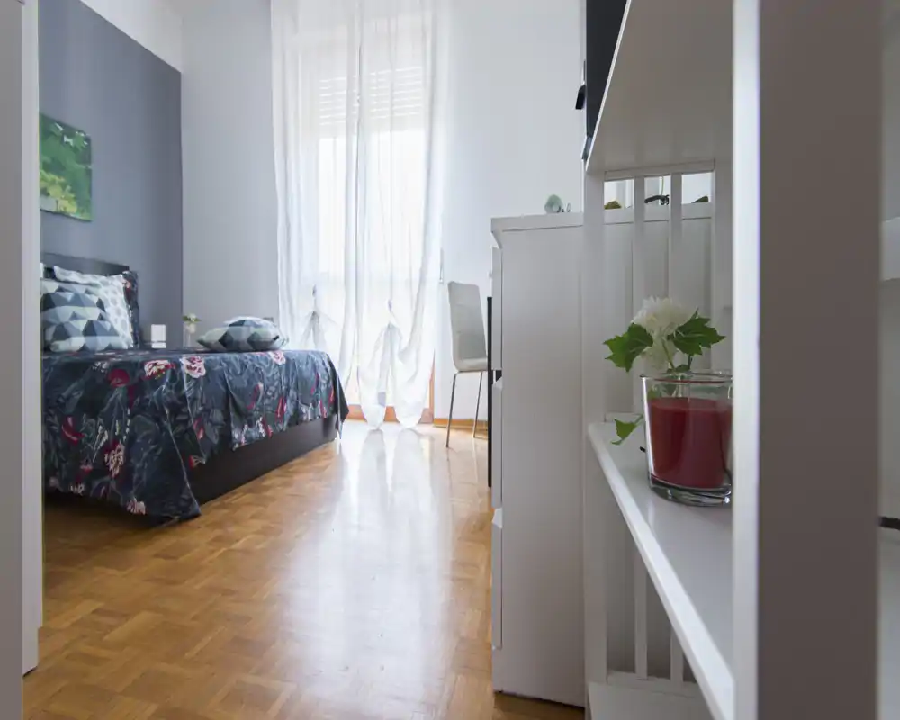 Camera da letto luminosa e arredata con letto matrimoniale con copriletto fantasia, finestra con tenda leggera, pavimento in parquet e una scrivania sullo sfondo — accogliente e ben illuminata.